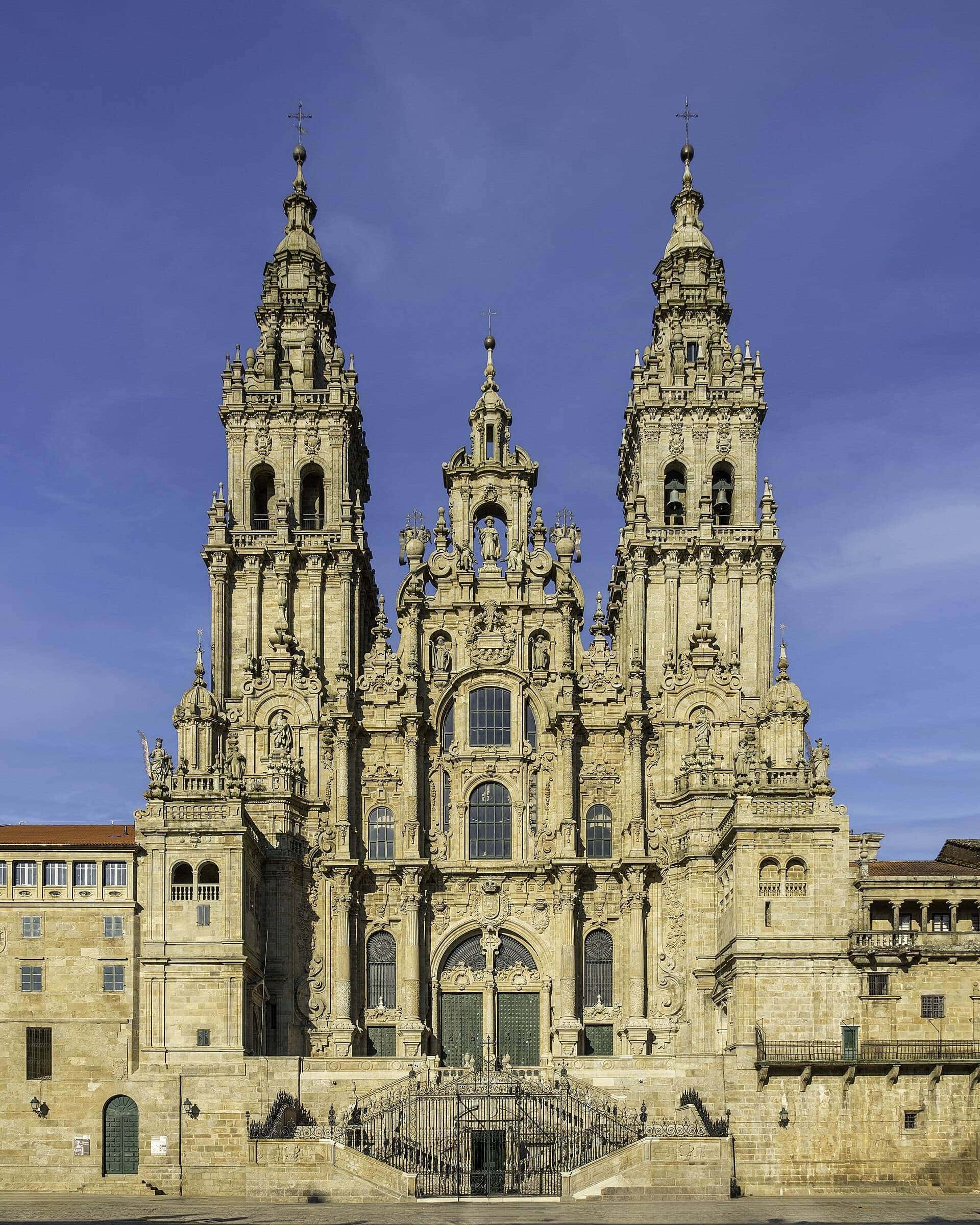 Fachada barroca del Obradoiro de la Catedral de Santiago, de frente y entera, con las dos torres gemelas, la escalinata de doble tramo y la verja de hierro al pie, bajo un cielo azul y sin nadie en la plaza